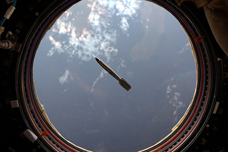 The half-scale Mini Hugo rocket, floating in front of a porthole window in the ISS. The earth is seen in the background through the window.